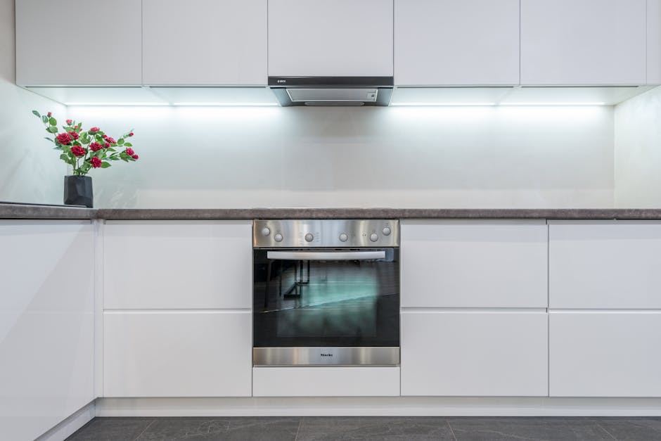 A modern kitchen with a large window above a beige countertop that has a double stainless steel sink and is cluttered with dishes, utensils, and cleaning items. Behind the sink, there are several bott